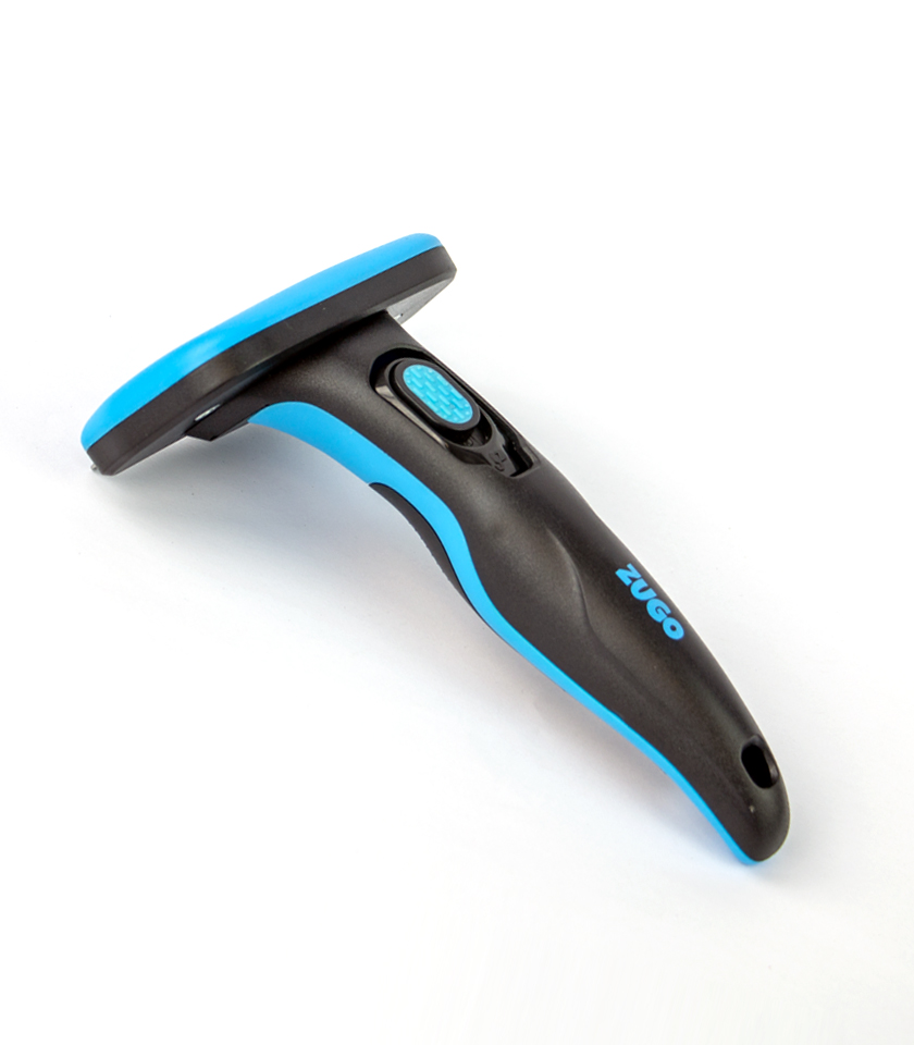 An angled studio shot of a black and blue Zugo Self Cleaning Deshedding Dog Brush. The brush has an ergonomic black handle with a blue accent, a blue button for the self-cleaning feature, and the word "ZUGO" printed on the side. The deshedding blade is not visible from this angle.