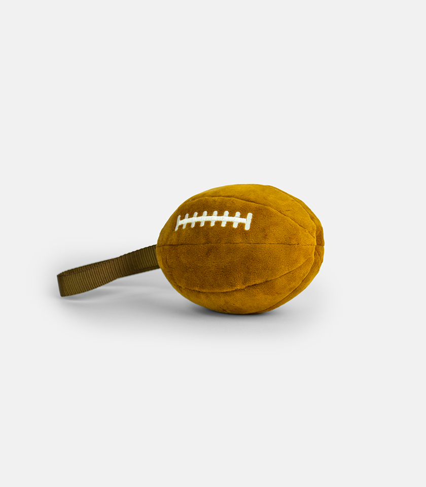 Plushball Plush Dog Toy, a brown plush toy shaped like a football with white stitching on top and a fabric loop handle on the side.