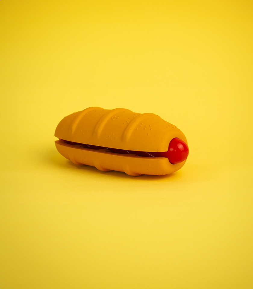 Hot Doggy Rubber Dog Toy, a hot dog-shaped toy with a textured orange bun and a red sausage.