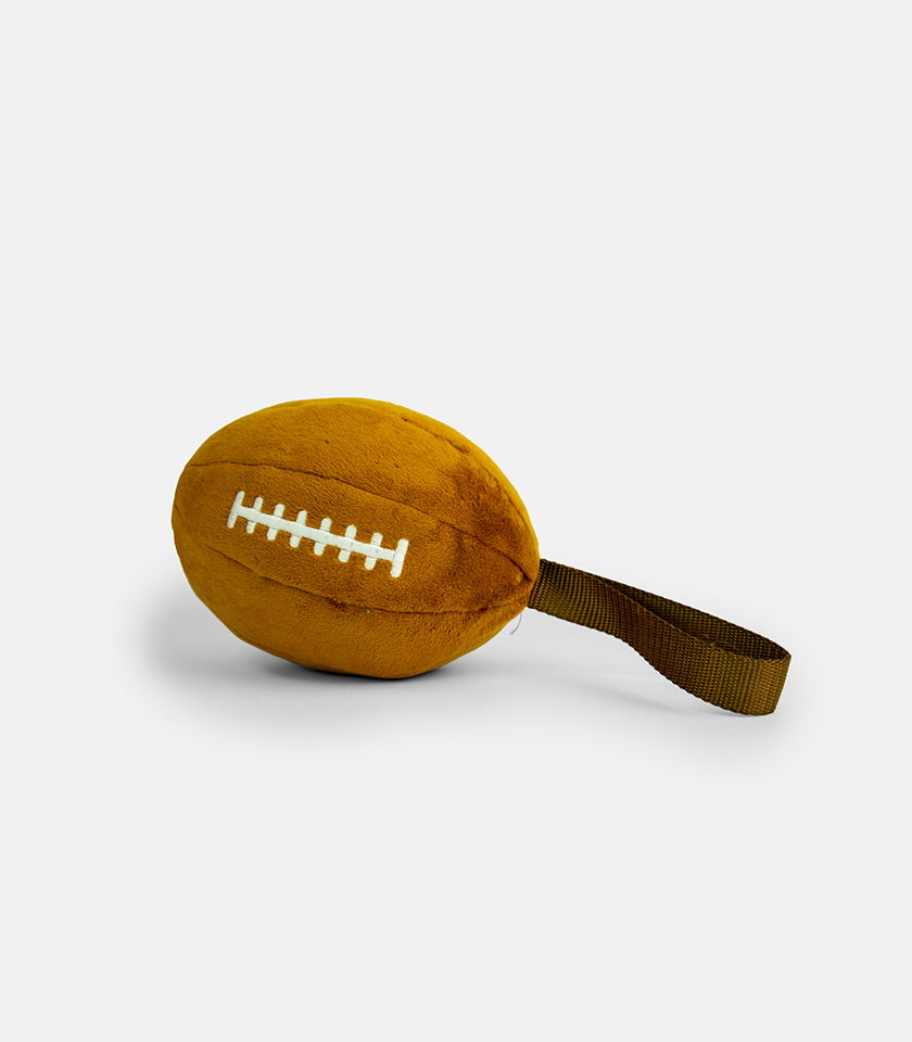 A close-up studio shot of a brown, plush Plushball Dog Toy shaped like an American football with white stitched laces. The toy has a dark green woven strap extending from one end, and is set against a solid light gray background.