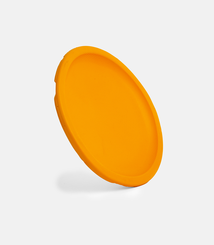 A close-up, angled view of a plain, solid orange Aeroglide EVA Dog Toy (flying disc) tilted almost vertically against a plain light gray background. The textured rim and flat surface of the disc are visible.