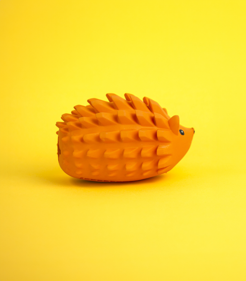 A solid orange rubber dog toy shaped like a hedgehog, with pronounced spikes along its back and a textured, rounded body. The small face, featuring a black eye and nose, is visible on the right, and the toy is set against a bright yellow background.